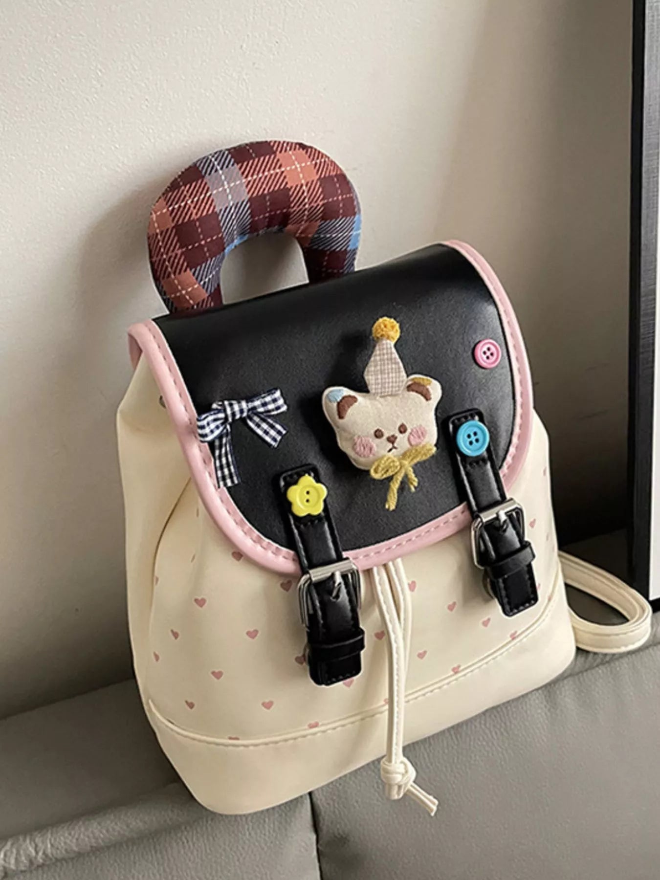 Beige backpack close-up: bear patch on front flap, plaid handle, heart print body