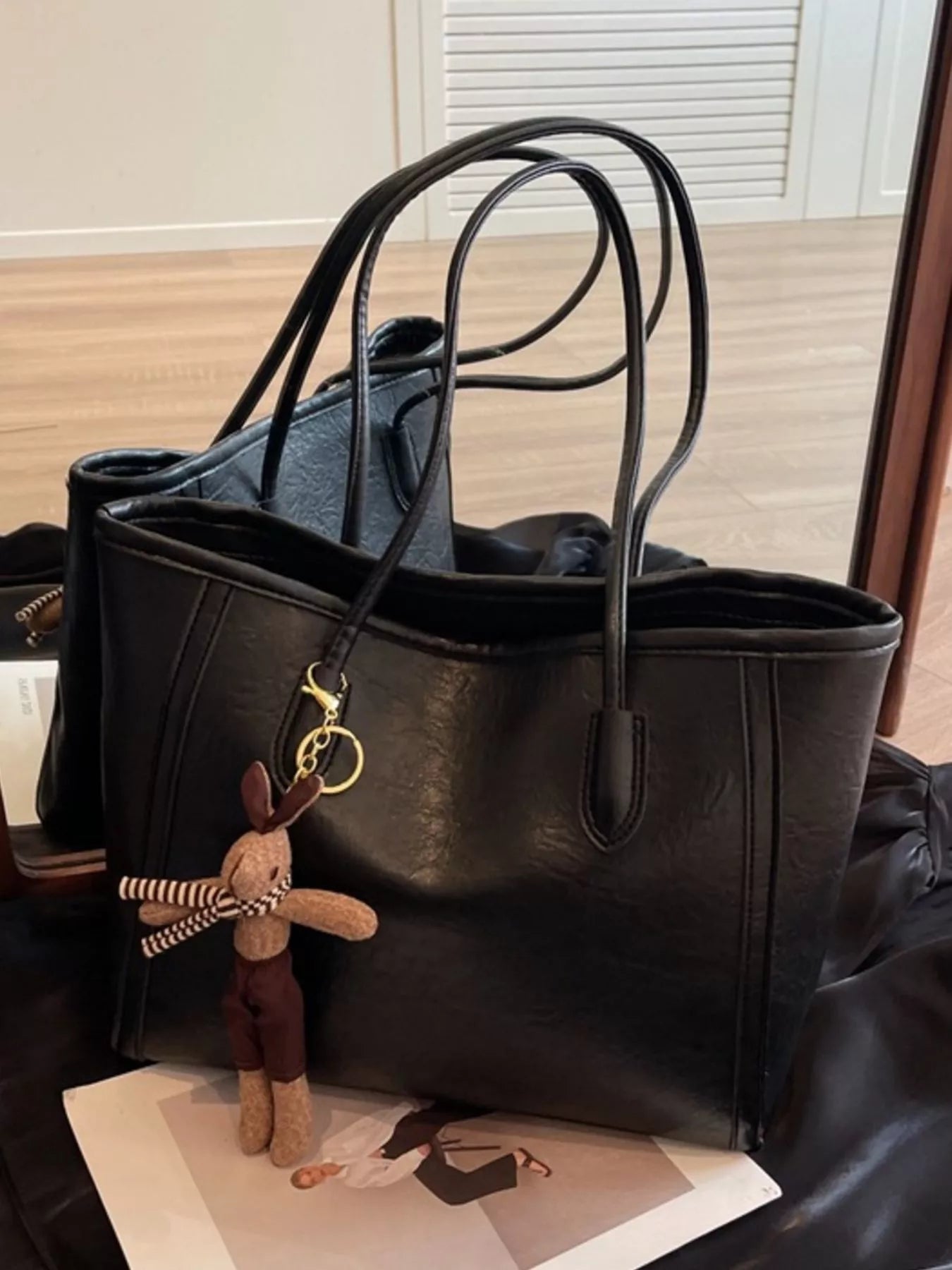 Black leather tote bag viewed from side, wide base, long handles, plush charm attached