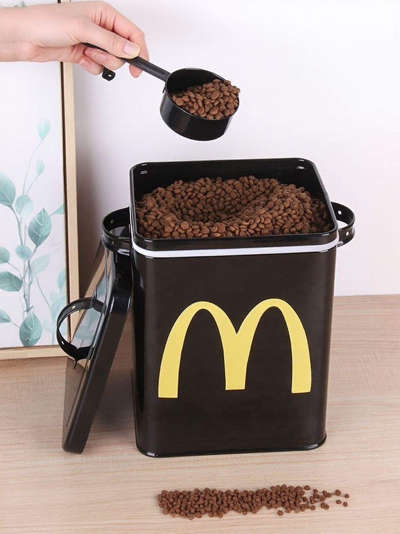 Black pet food storage bucket with handle, yellow logo, and kibble being poured in