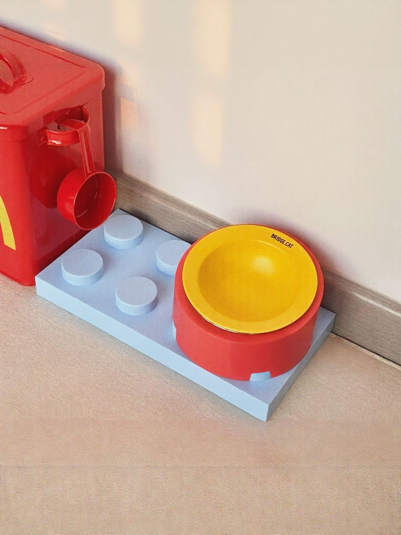 Red pet food storage bucket corner with side dispenser and yellow bowl on brick base