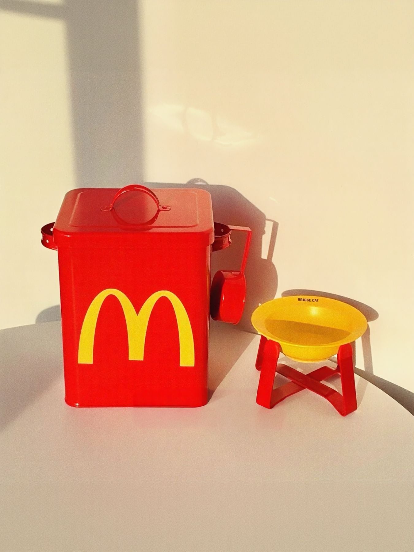 Red pet food storage bucket with handle and yellow logo beside yellow feeding bowl